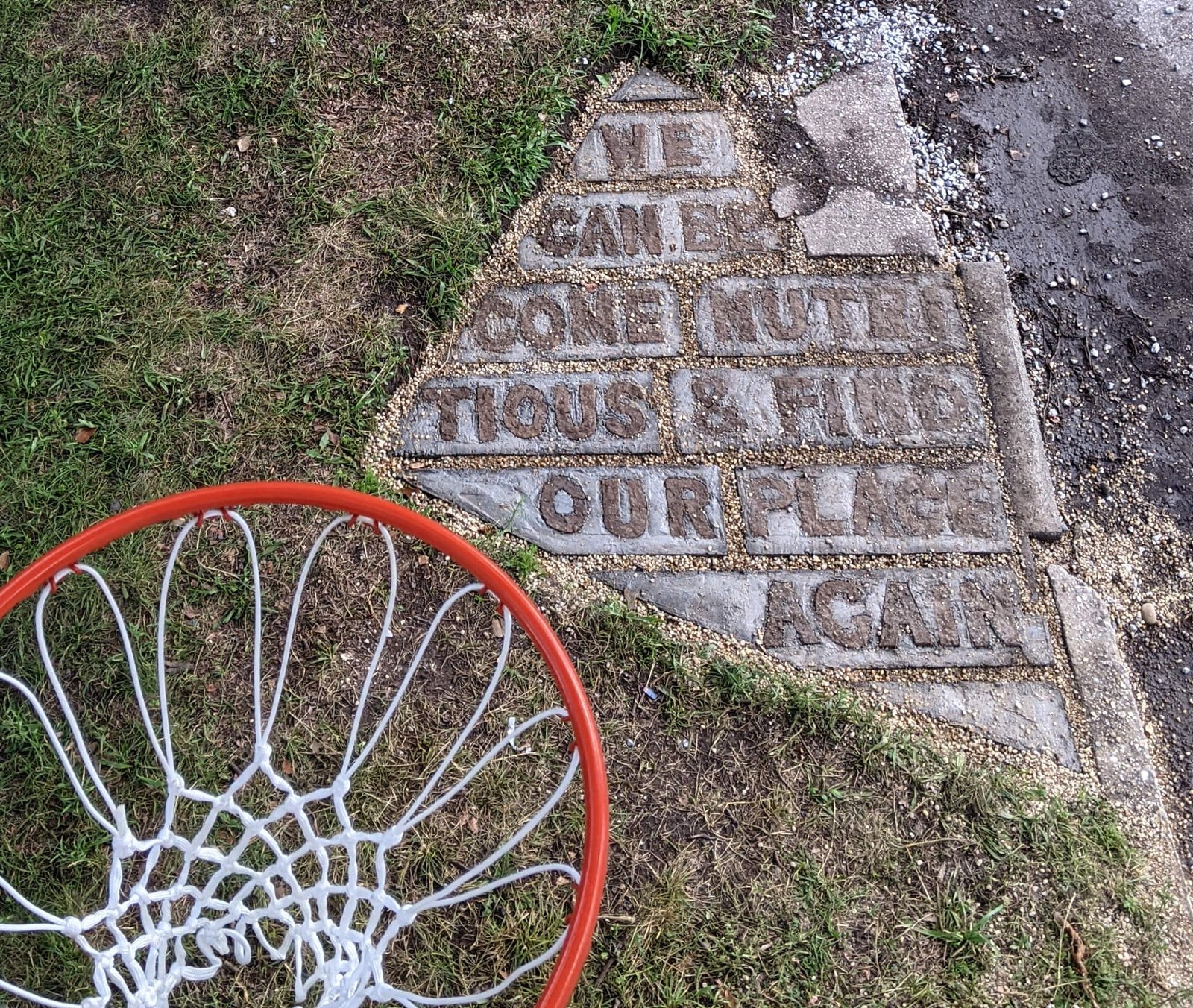 3 people play basketball on a concrete free throw lane that has text carved into it. A small house holds two more basketballs. A garden and a purple martin house.